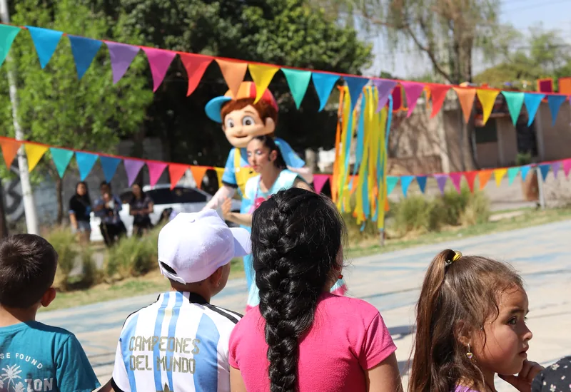 Niños y niñas jugando en el festejo del Día del Niño, con banderines de colores en la canchita.